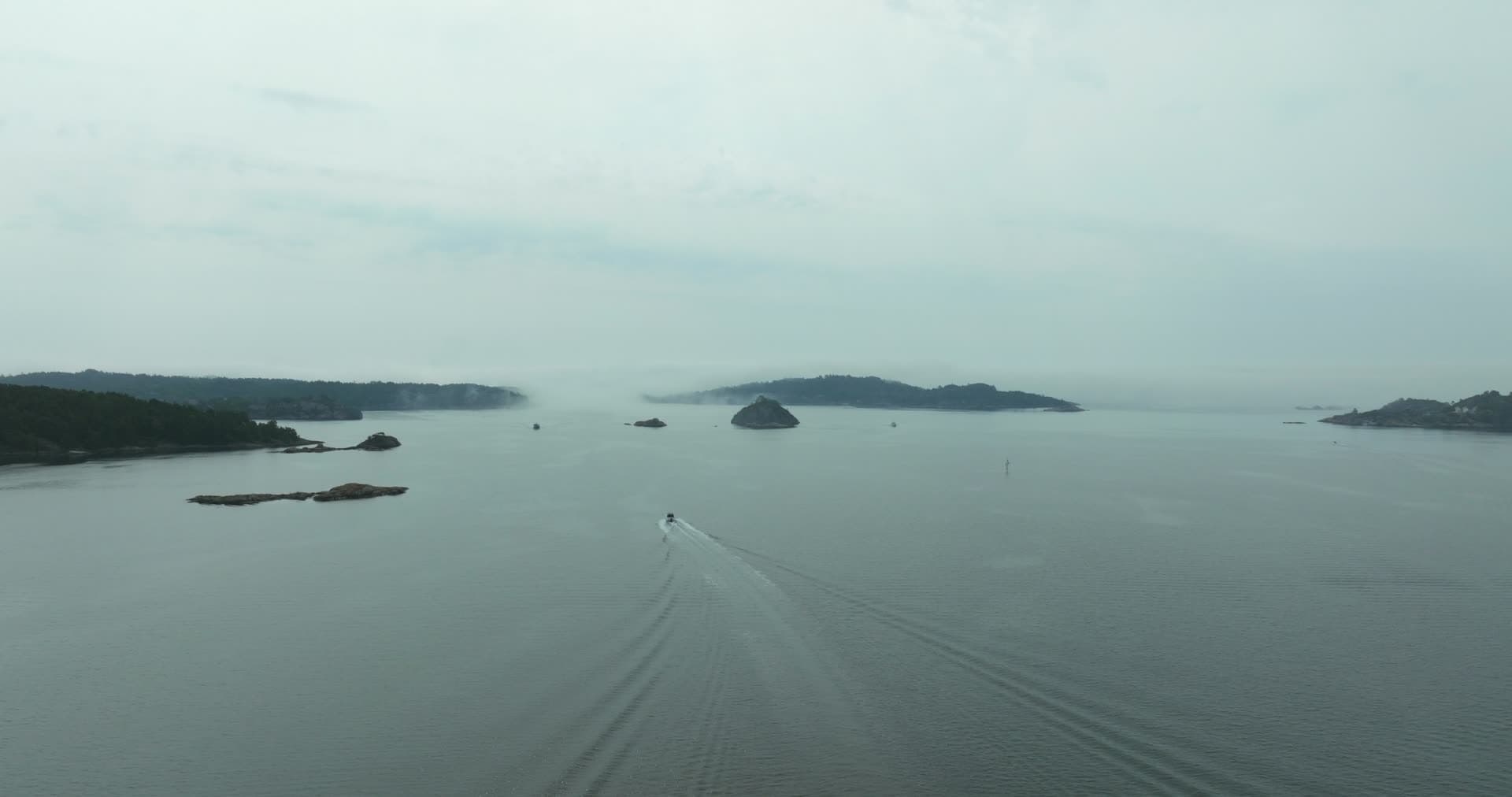 Motorboat trailing wake past low skerries in mist, Høllen, Norway
