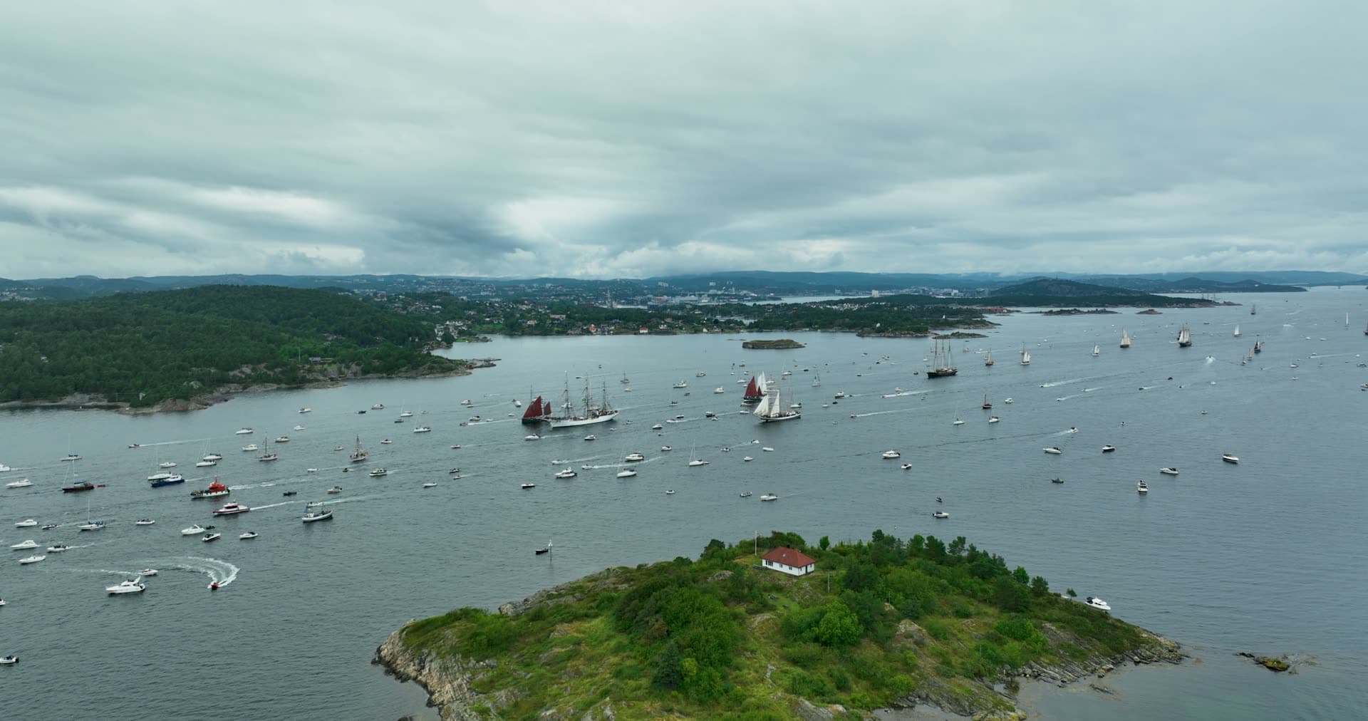 Tall ships parade past small red-roofed islet, Kristiansand, Norway