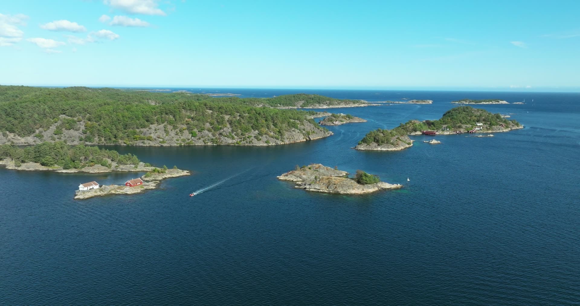 Small boat passing skerries with red boathouse, Lillesand, Norway
