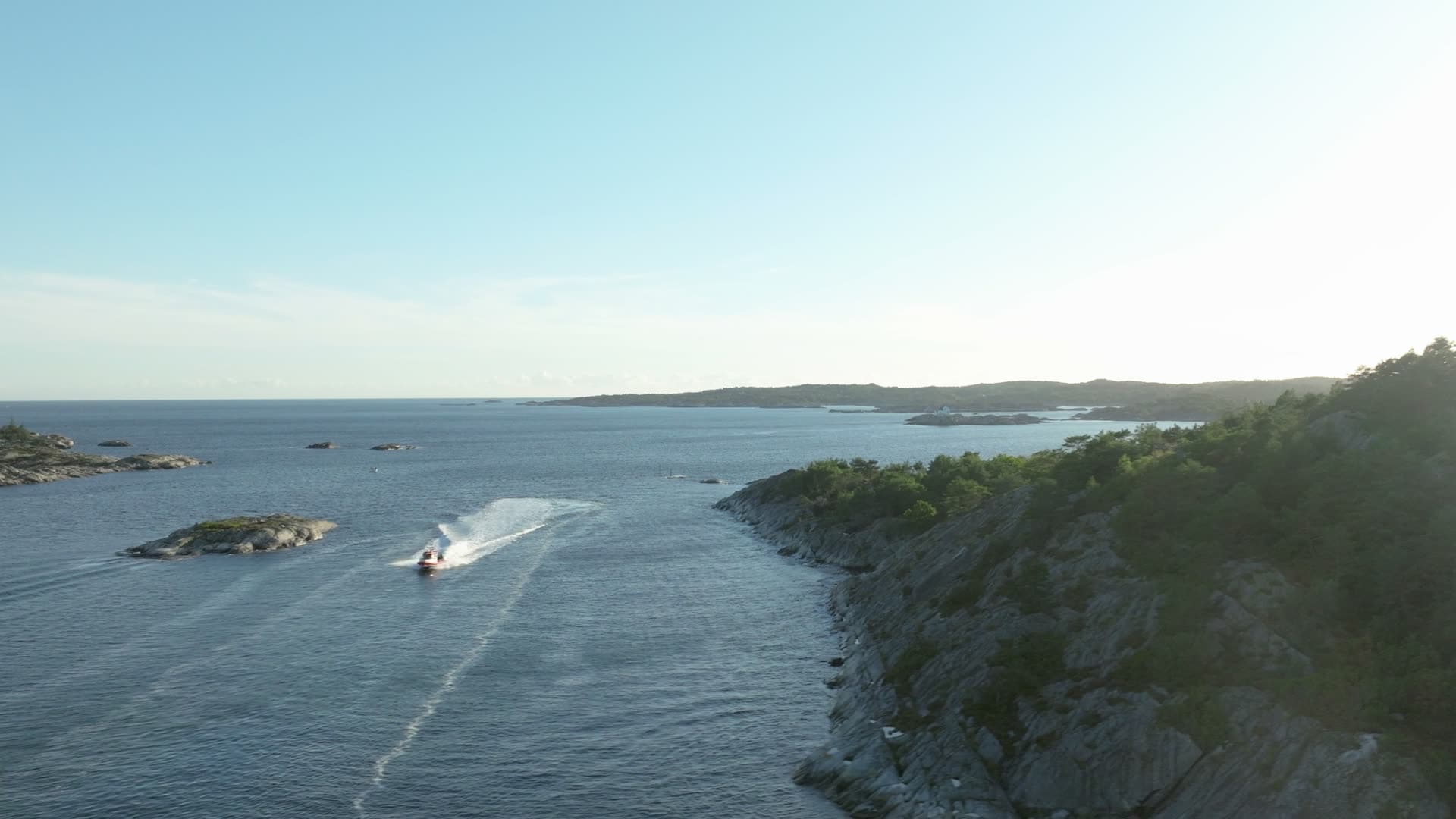 Motorboat wake past cliff into low sun, Lillesand, Norway