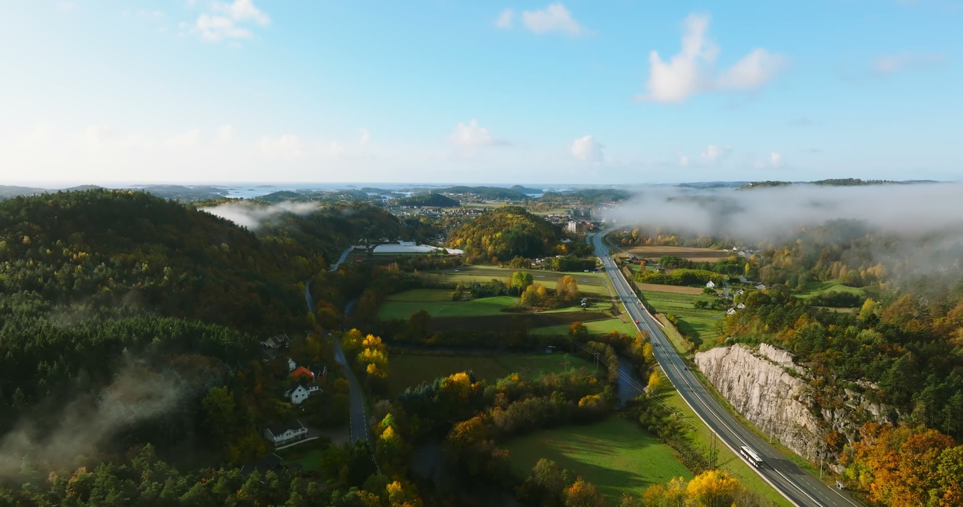 Highway past rock cut, fog over farmland, Monan, Norway
